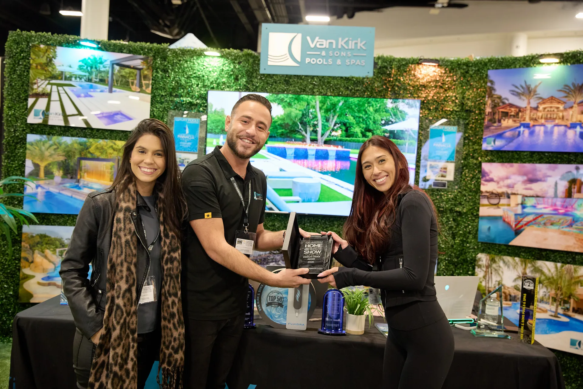 Pool company booth staff chatting with a show host, pool imagery on TV screens behind them