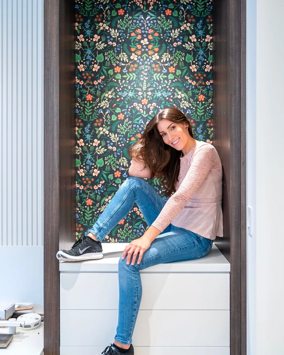 Katie Hunt seated in front of a dark floral feature wall in a white built-in nook.